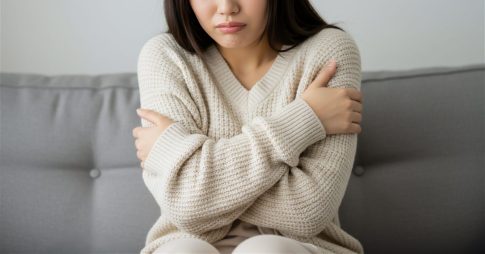 寒さに耐える若い日本人女性のイメージ。クリーム色のニットセーターを着て、腕を抱えるようにして寒そうな仕草をしている。顔は一部が見えるだけで、寒さによる不快感が表情から伝わる。背景はソファとぼんやりとしたニュートラルな色合いで、冷え性の雰囲気を強調している。